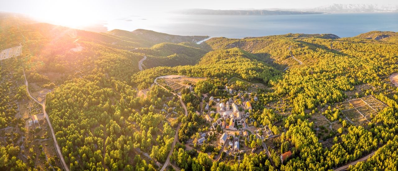 An aerial view of Humac on the island of Hvar, a small settlement on a hilly island surrounded by dense wooded areas and winding roads.