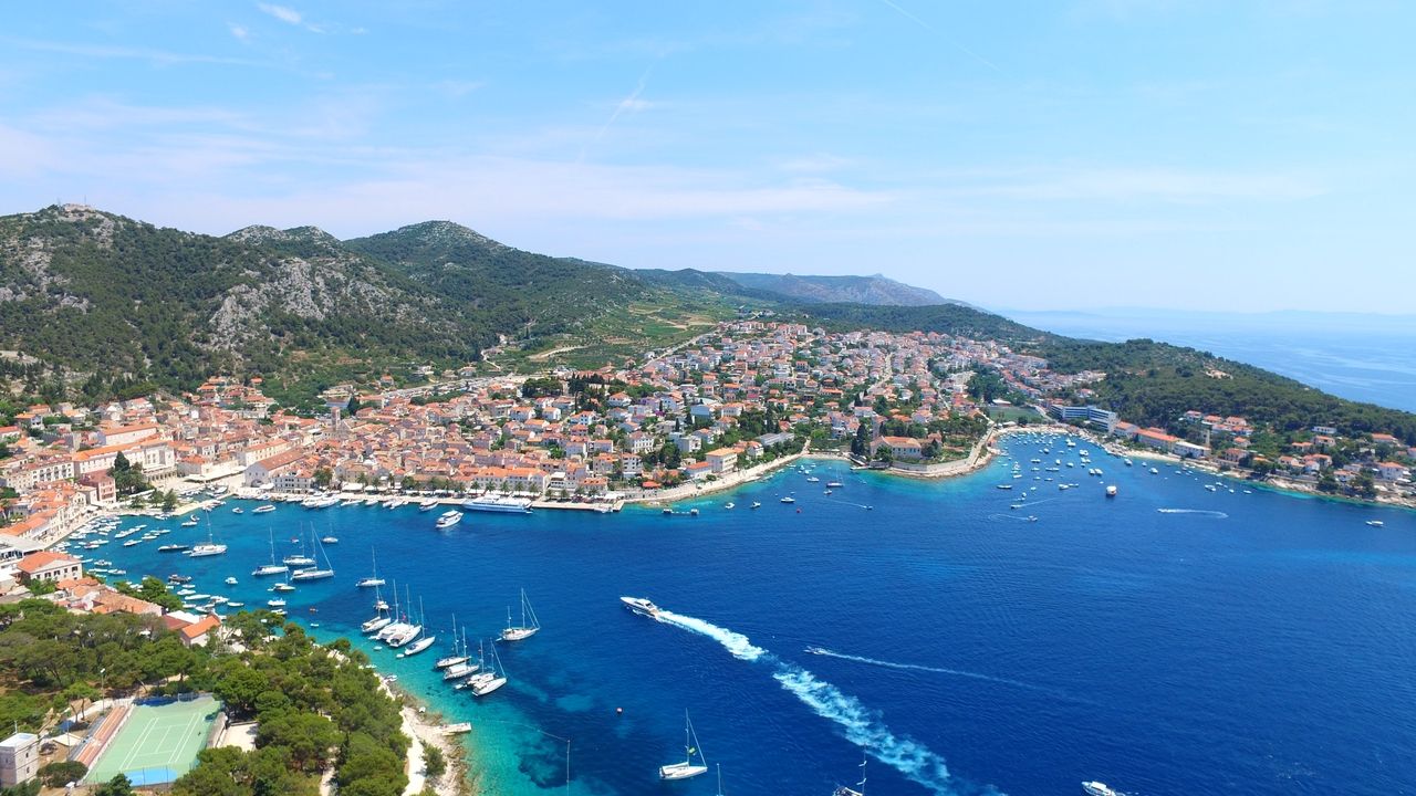 O vedere aeriană a orașului Hvar pe insula Hvar. Orașul se află pe coastă, cu numeroase bărci și iahturi în apa albastru închis a portului. Acoperișurile roșii ale clădirilor se întind pe deal, înconjurate de dealuri verzi.