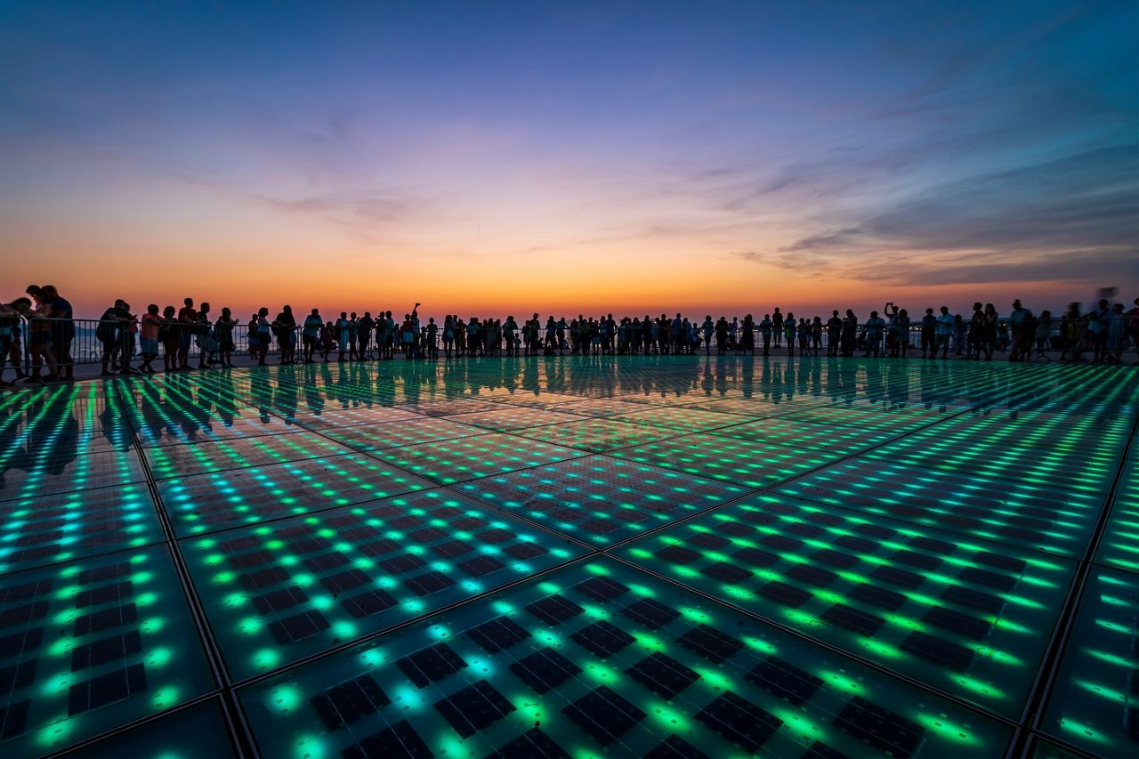 The "Greeting to the Sun" shows a large circular installation with colored LED lights in the ground. Many people stand around the installation enjoying the light show.