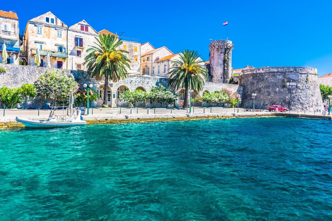 Costa de Korčula con edificios históricos y una impresionante muralla de piedra al fondo. El paseo marítimo está bordeado de palmeras y plantas florecientes. En primer plano, el agua cristalina y azul del Adriático brilla.