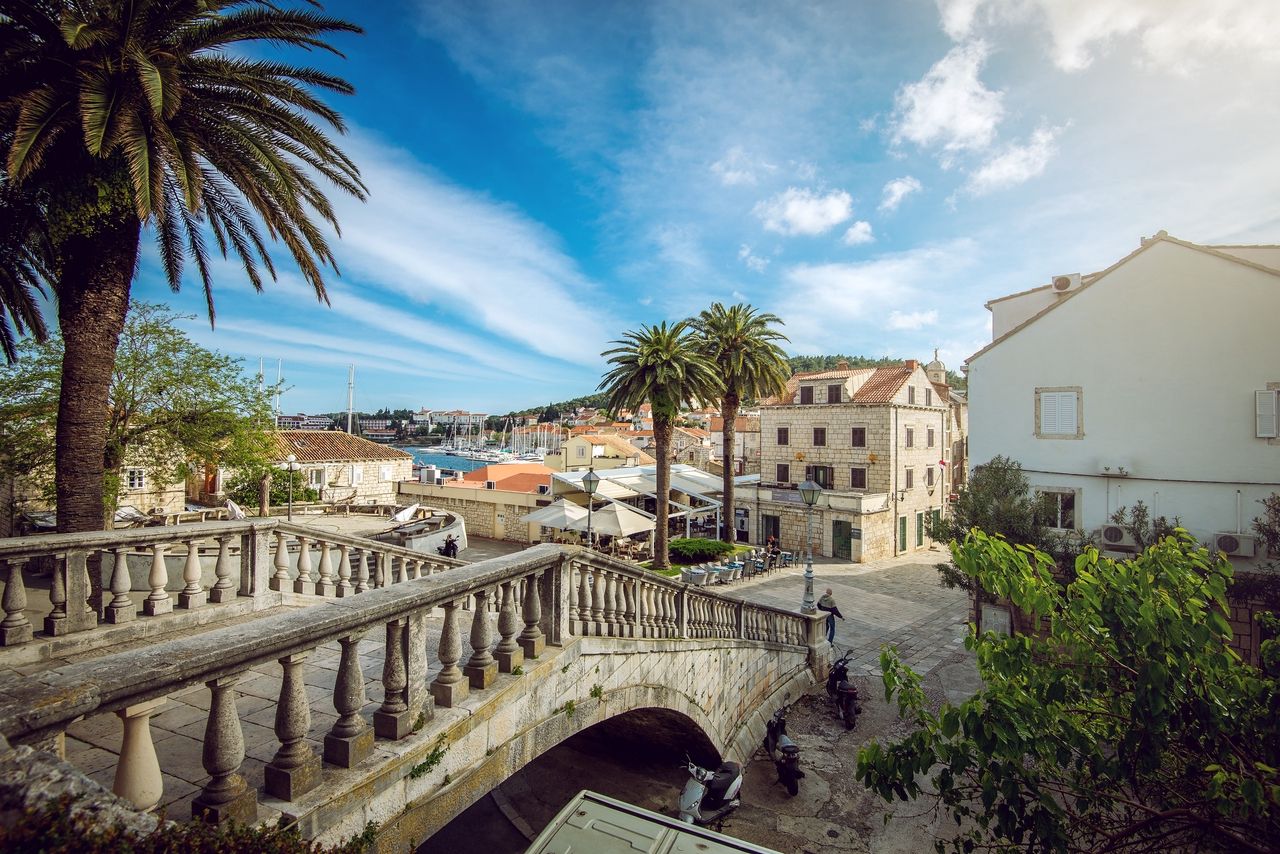 A stone bridge in Korčula leading over a cobbled street. In the background, historic buildings, palm trees, and the marina can be seen.