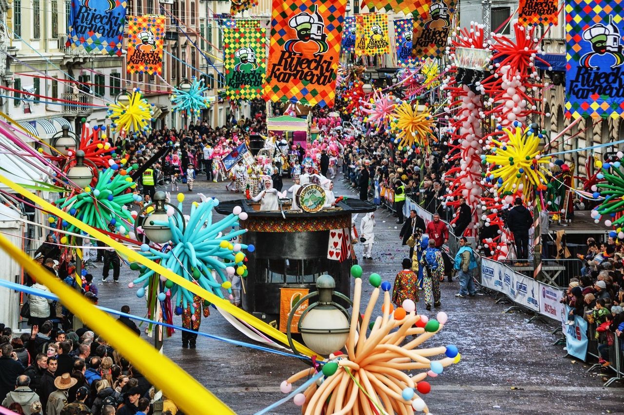 Carnival in Rijeka, with colorful balloon decorations and floats parading through a busy street. Crowds line the streets celebrating the festival.