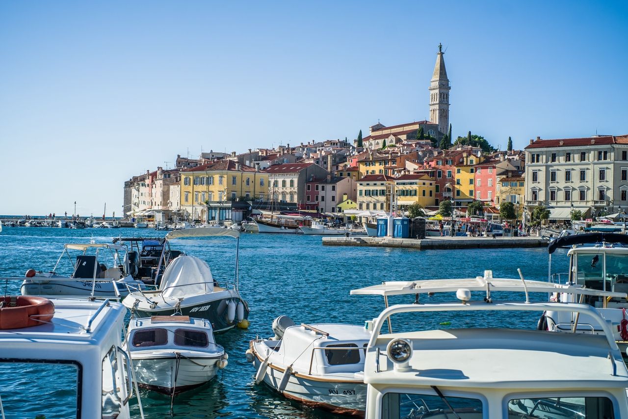 Havnen i Rijeka, med mange båter i forgrunnen som flyter i det klare blå vannet. I bakgrunnen er den historiske gamlebyen i Rijeka synlig, med fargerike hus og den markante kirken som troner på en høyde.