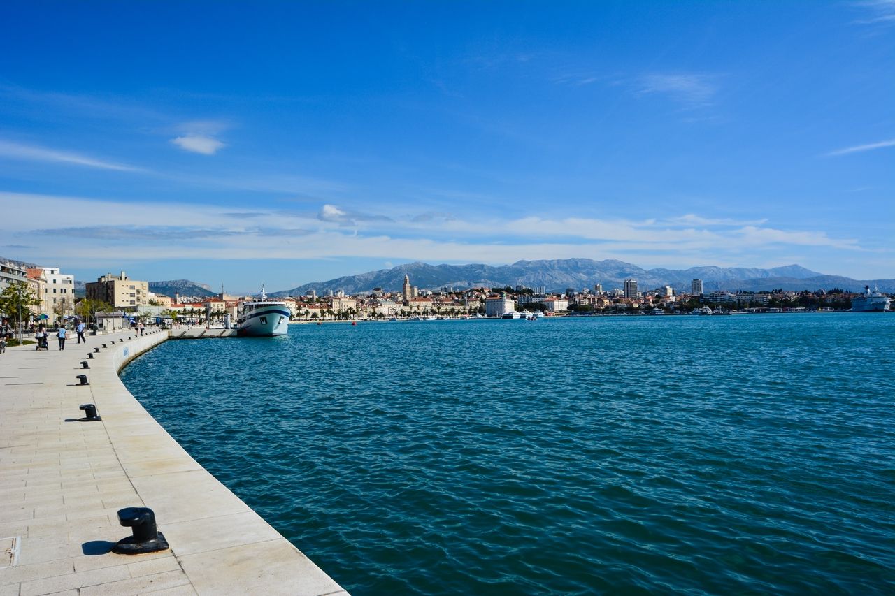 Une vue large sur la promenade de Split avec un grand navire amarré dans le port. La promenade est animée de promeneurs et longe l'eau bleu profond. En arrière-plan, la ville historique de Split et les montagnes environnantes sous un ciel bleu clair.