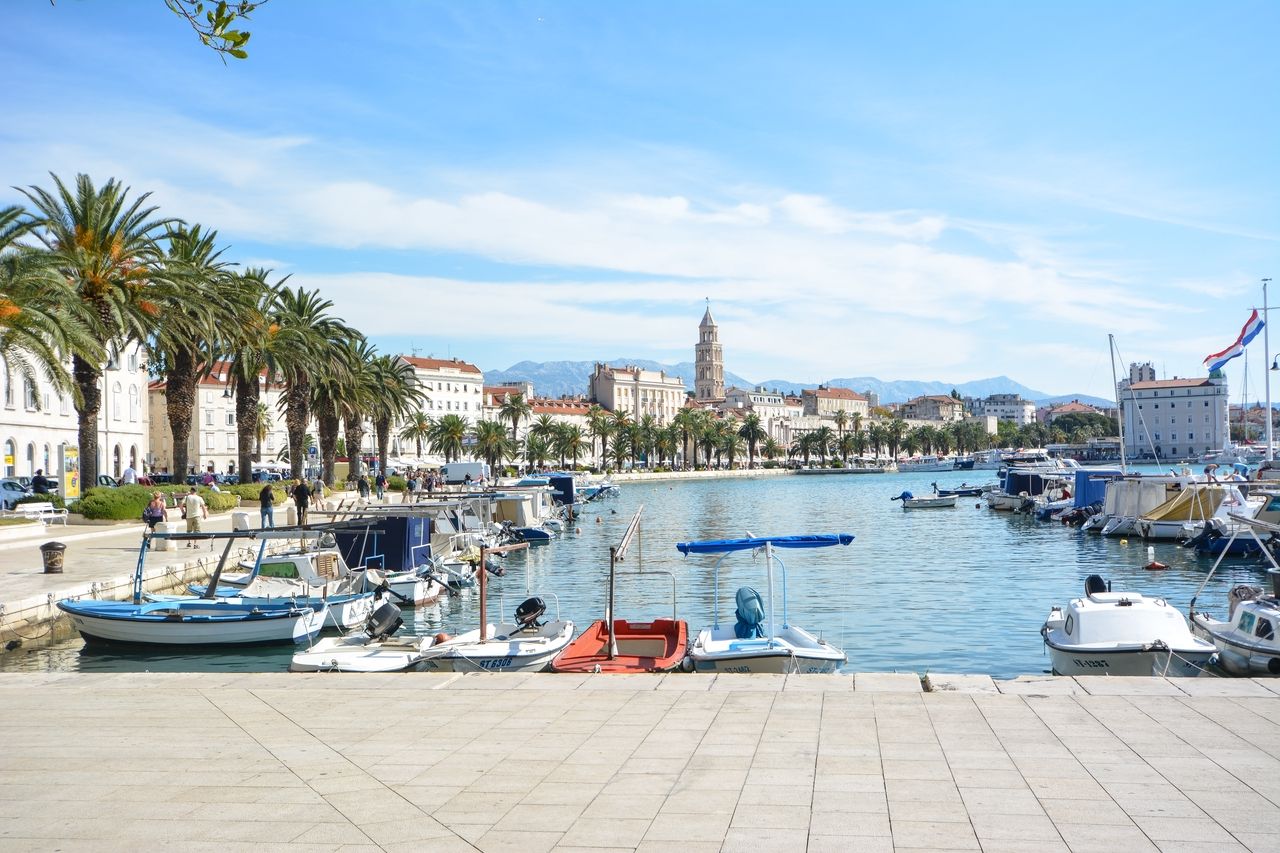 A picturesque view of the waterfront promenade in Split, lined with tall palm trees and historic buildings. In the foreground, several boats can be seen in the calm water of the harbor. In the background, the bell tower of Diocletian's Palace rises, and the surrounding mountains provide an impressive backdrop.