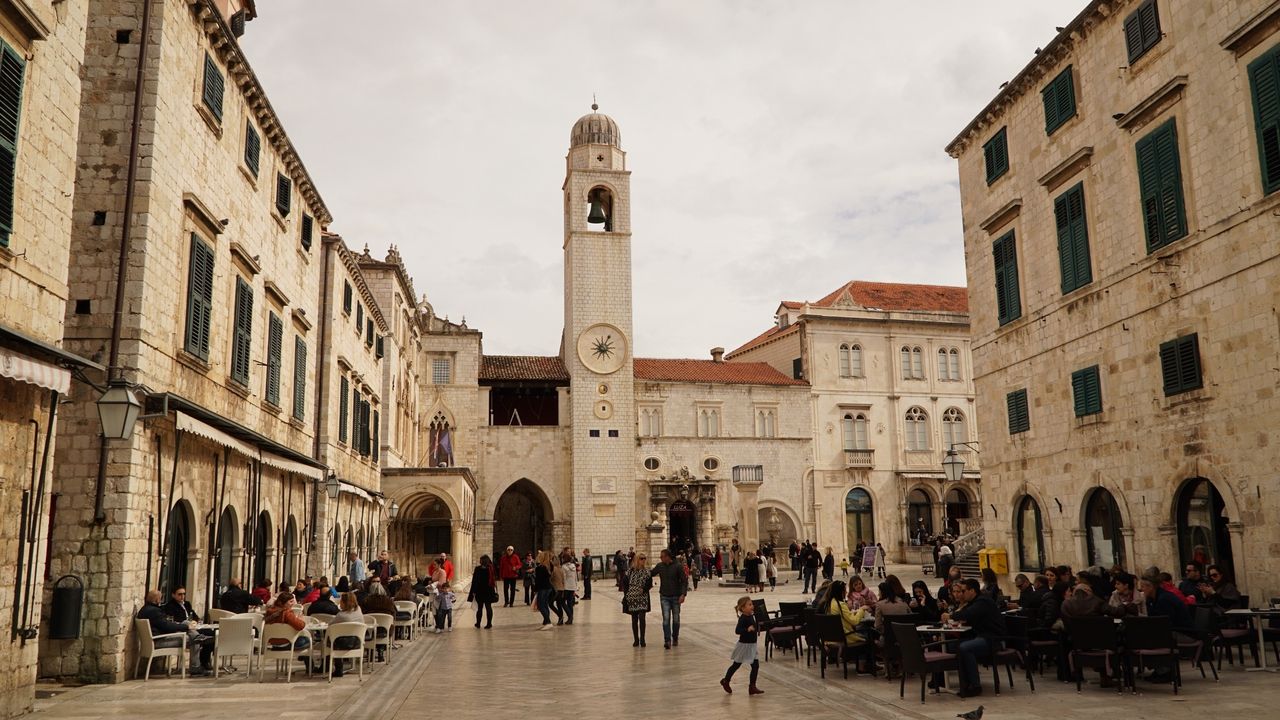 Plass foran klokketårnet og Sponza-palasset i Dubrovnik, med mennesker på kafeer