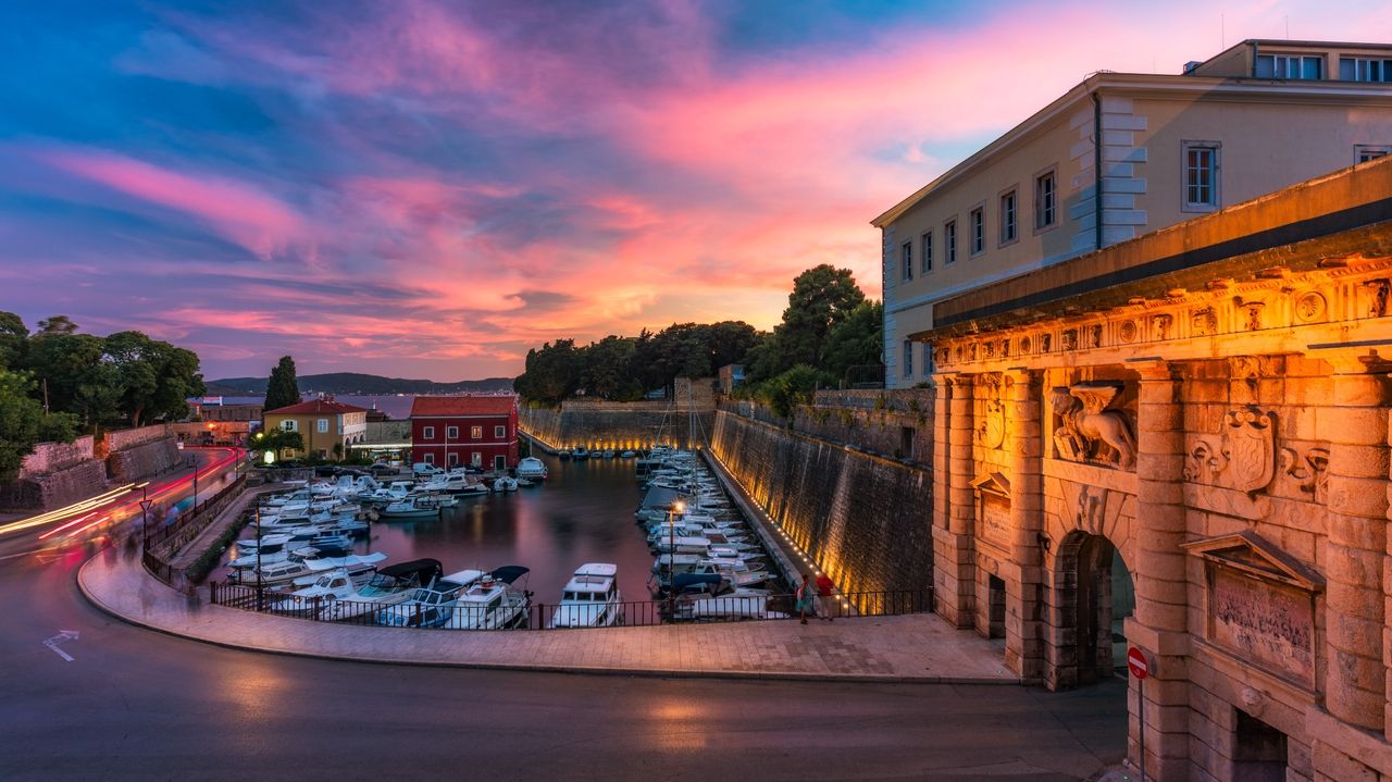 Zadar auringonlaskun aikaan, pieni satama täynnä veneitä, jota ympäröivät historialliset muurit ja rakennukset. Kaupungin lämpimät valot heijastuvat vedestä, kun taivas hehkuu vaaleanpunaisen ja violetin sävyissä. Etualalla näkyy historiallinen portti, joka on valaistu oranssilla valolla.