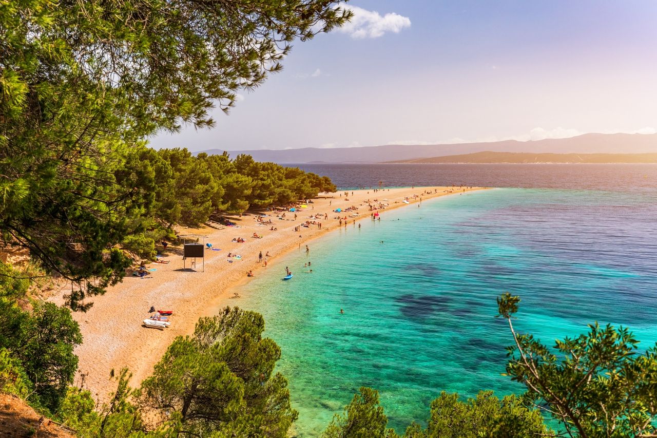 Pogled na živahnu plažu Zlatni rat na otoku Braču. Plaža je okružena bujnim borovima i proteže se u uskom poluotoku u kristalno čistu, tirkiznu vodu.