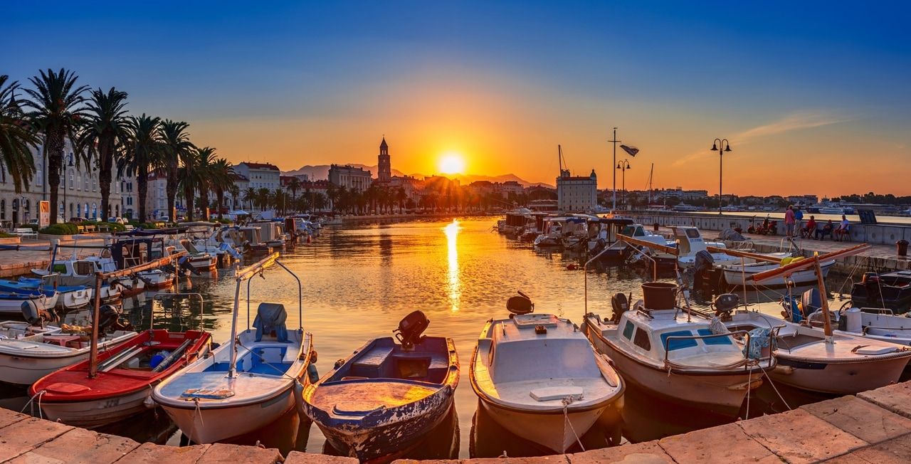 A naplemente a festői split-i kikötő sétány felett Horvátországban, pálmafákkal és számos hajóval, amelyek békésen fekszenek a vízen. A háttérben történelmi épületek és egy templom látható, amelyek kiemelik a város kulturális szépségét.