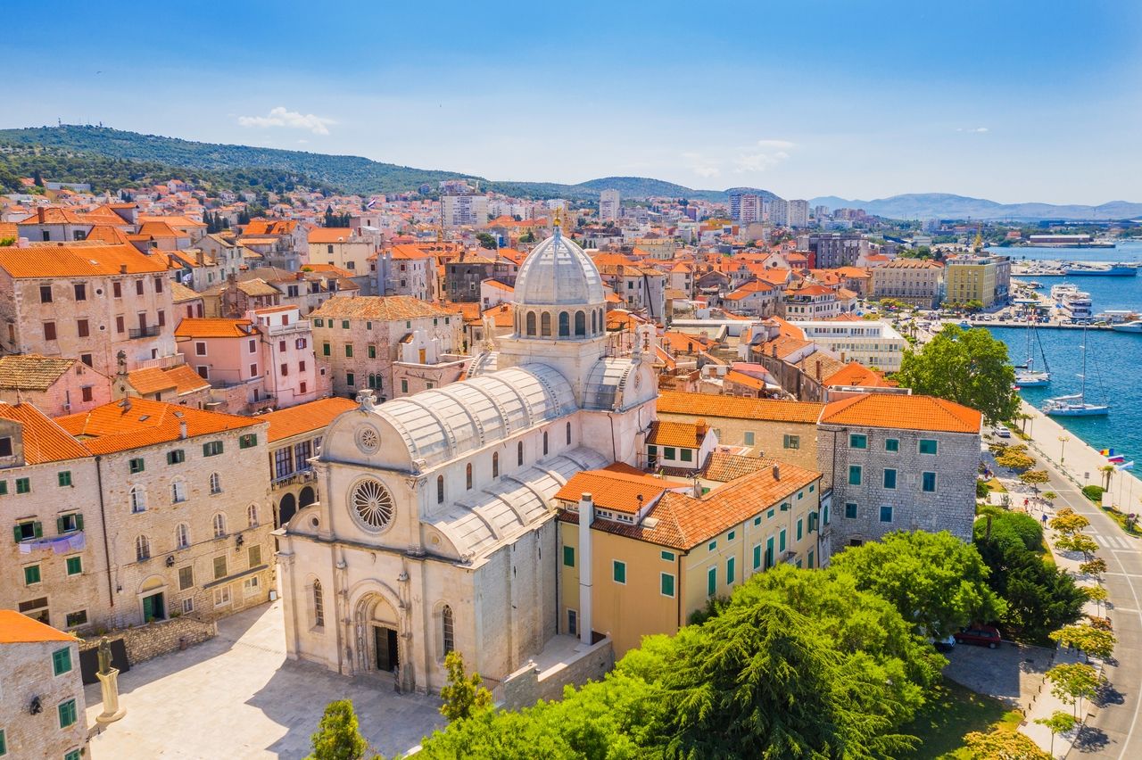 Vista aérea de Šibenik con la catedral distintiva en primer plano. La ciudad se caracteriza por sus techos rojos tradicionales y edificios históricos de piedra. En el fondo se ven edificios modernos y el pintoresco puerto con veleros.