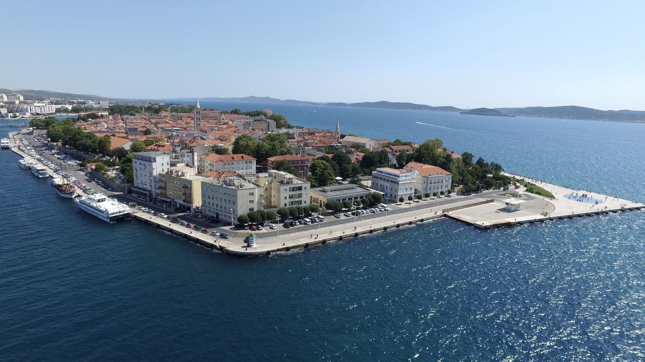 Vista aérea de la ciudad costera de Zadar, mostrando el casco antiguo con sus edificios históricos y techos rojos. A lo largo del paseo marítimo se ven numerosos barcos y barcos. En primer plano se encuentra la plaza con el famoso Órgano del Mar y el Saludo al Sol.