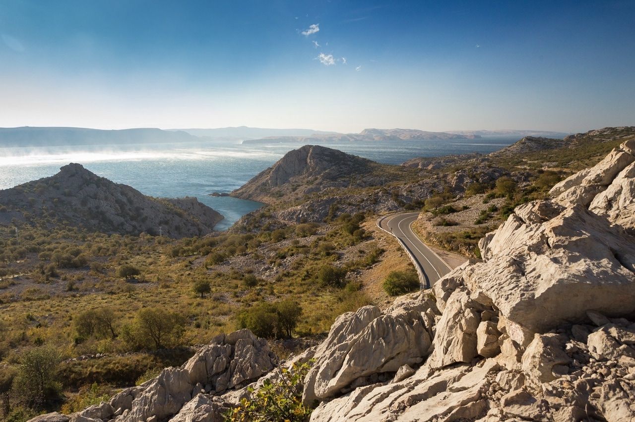 Panoramablick auf eine kurvenreiche Straße entlang der Küste Kroatiens, die sich durch die felsige Landschaft schlängelt. Im Hintergrund glitzert das Adriatische Meer unter dem klaren, blauen Himmel.