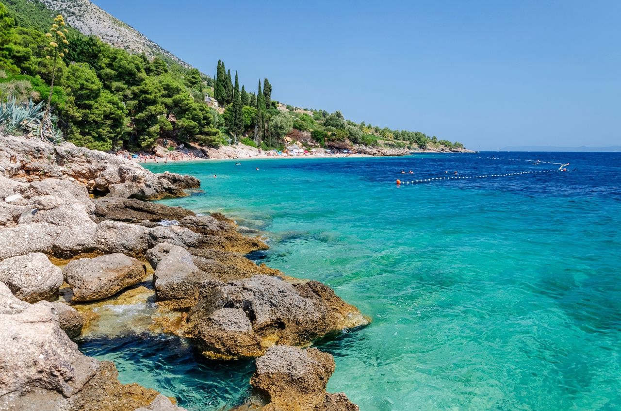 Malowniczy krajobraz wybrzeża na wyspie Brač z krystalicznie czystą, turkusową wodą i skalistym wybrzeżem. W tle widać idylliczną plażę z kąpiącymi się ludźmi, otoczoną bujną roślinnością i lasami sosnowymi.