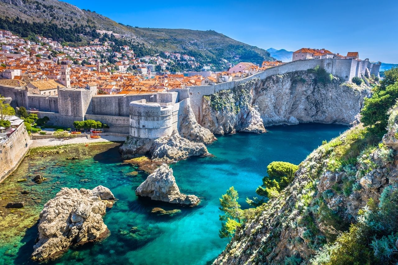 Aerial view of the old town of Dubrovnik in Croatia. The medieval city walls surround the city, which sits on a cliff above the azure sea. The red tiled roofs of the buildings contrast with the clear, blue water. In the background, the green hills that surround the city can be seen.