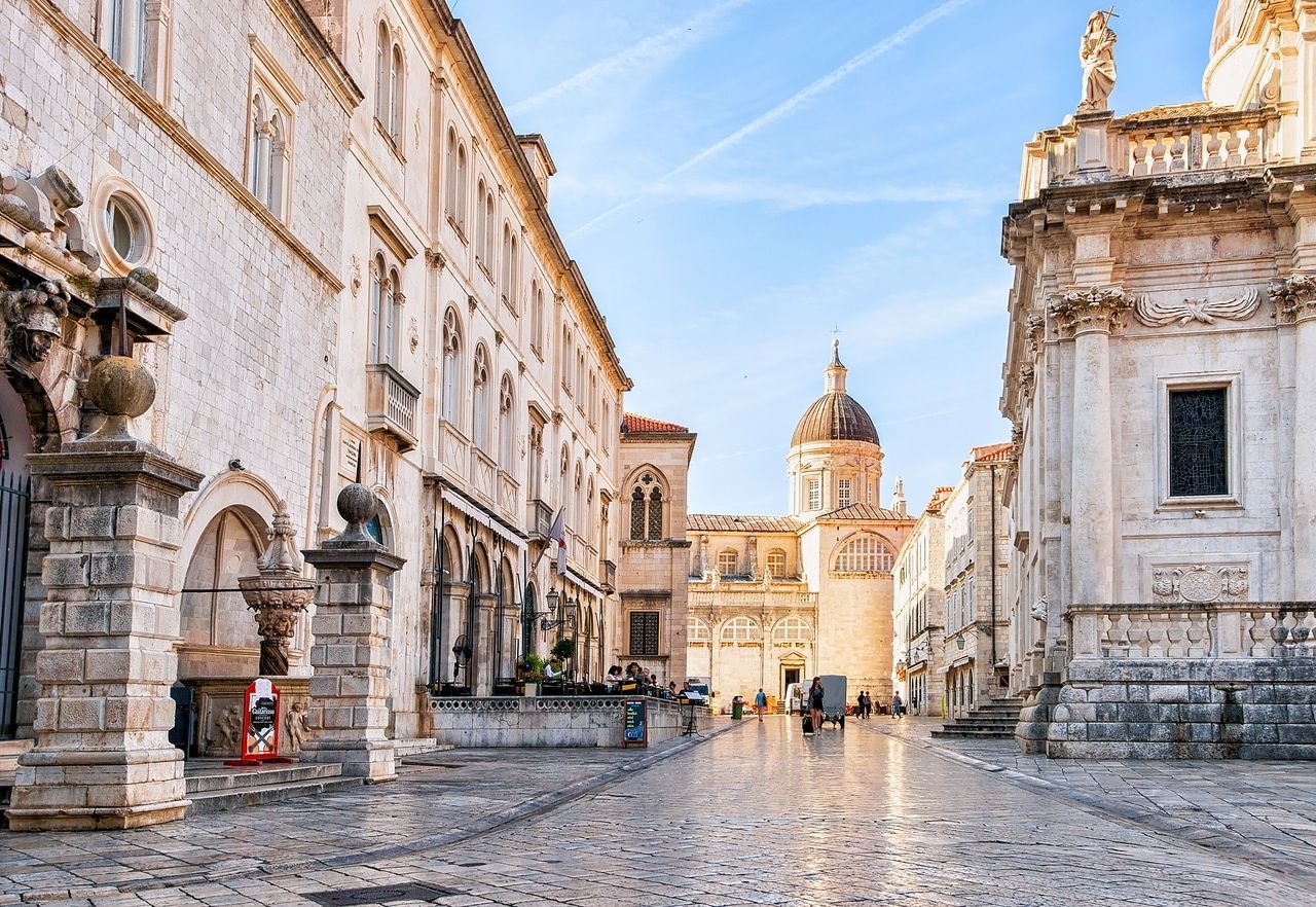 Egy fotó Dubrovnik egyik macskaköves utcájáról, amelyet világos kőből készült történelmi épületek vesznek körül. A háttérben egy jellegzetes kupolával rendelkező templom látható. Az épületek homlokzata dekoratív elemekkel és ablakokkal van díszítve. Néhány ember sétál az utcán, miközben az ég enyhén felhős.