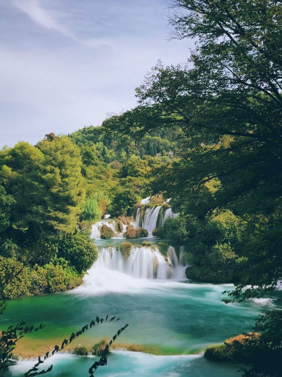 Waterval te midden van een groen boslandschap. Een schilderachtige waterval, omgeven door weelderige, groene bomen en planten. Het water stroomt zachtjes in een helder, turquoise bassin. De lucht is licht bewolkt en de dichte vegetatie creëert een rustige en natuurverbonden sfeer.