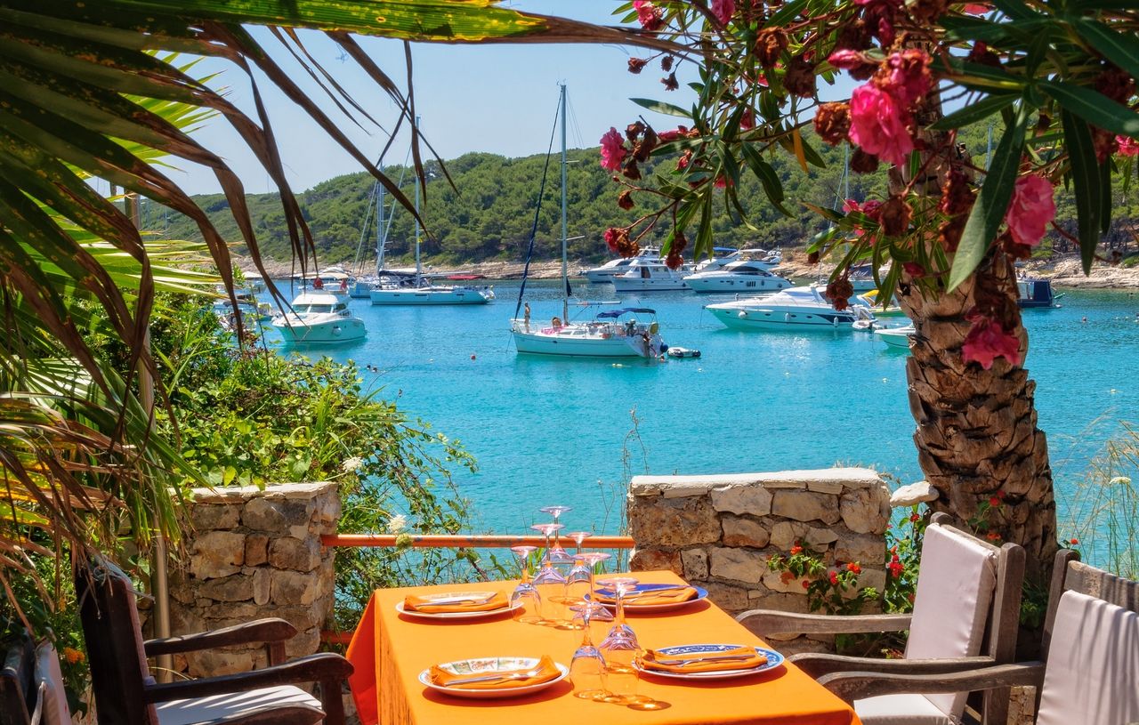 Un arrangement de repas en plein air idyllique avec vue sur une magnifique baie aux eaux turquoise et de nombreux bateaux. La table est dressée avec une nappe orange et entourée de verdure luxuriante et de plantes en fleurs.