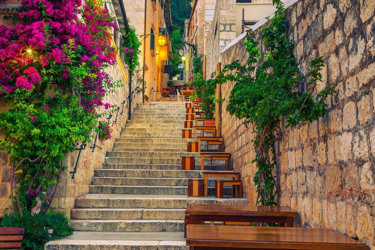 Una empinada escalera de piedra en Hvar, Croacia, flanqueada por flores coloridas y vegetación exuberante. A lo largo de la escalera hay pequeñas mesas y bancos de madera que crean una atmósfera acogedora. Las paredes de los edificios son de piedra clara y en el fondo se ven faroles que emiten una luz cálida. La escena irradia un ambiente encantador y acogedor.