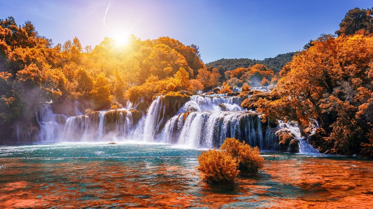 Die Wasserfälle im Nationalpark Krka in Kroatien im Herbst. Das Wasser stürzt über mehrere Kaskaden in einen türkisfarbenen Fluss, umgeben von Bäumen in leuchtenden Herbstfarben. Die Sonne scheint hell und taucht die Szene in ein warmes, goldenes Licht, das die farbenfrohe Landschaft noch intensiver erscheinen lässt.