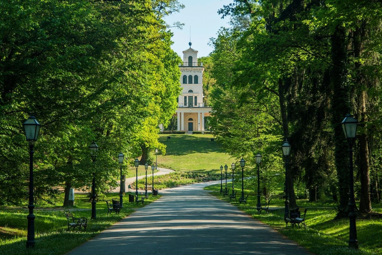 A path in Maksimir Park in Zagreb, lined with street lamps and benches surrounded by lush green foliage. At the end of the path stands a historic building with a bright facade and a tower. The scene conveys a calm and peaceful atmosphere, ideal for a walk on a sunny day. The park is well-maintained and offers a relaxing retreat amidst nature.