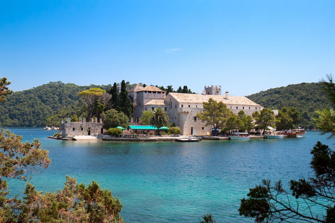 En pittoresk vy av Benediktinerklostret på ön Mljet, Kroatien. Det historiska klostret ligger på en liten ö mitt i en klar, turkos sjö. Omgivet av frodig grönska och blå himmel erbjuder klostret en lugn och idyllisk atmosfär. Båtar är förtöjda vid stranden, och några människor njuter av den fridfulla omgivningen.