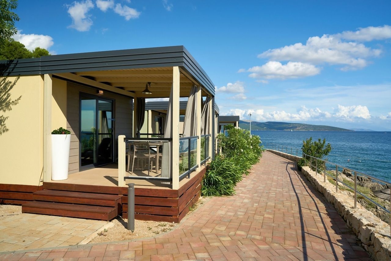 Modernes Strandbungalow mit Meerblick. Ein elegantes Bungalow mit überdachter Terrasse und großen Fenstern, die den Blick auf das blaue Meer freigeben. Ein gepflasterter Weg führt entlang der Küste und ist von grünen Pflanzen gesäumt. Der Himmel ist klar mit wenigen Wolken, was eine entspannte und sonnige Atmosphäre schafft.