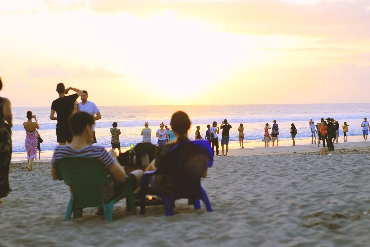 Människor njuter av en vacker solnedgång på en sandstrand. Några sitter på stolar och slappnar av, medan andra promenerar längs stranden och njuter av den fridfulla atmosfären och den varma kvällssolen.