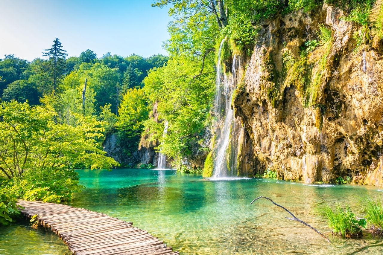 Parcul Național Lacurile Plitvice din Croația cu un traseu de drumeție din lemn care trece peste apă cristalină, de culoare turcoaz, înconjurat de vegetație verde luxuriantă. În fundal, mai multe cascade curg pe pereții de stâncă în lac. Parcul emană frumusețe naturală și o atmosferă liniștită, nealterată.