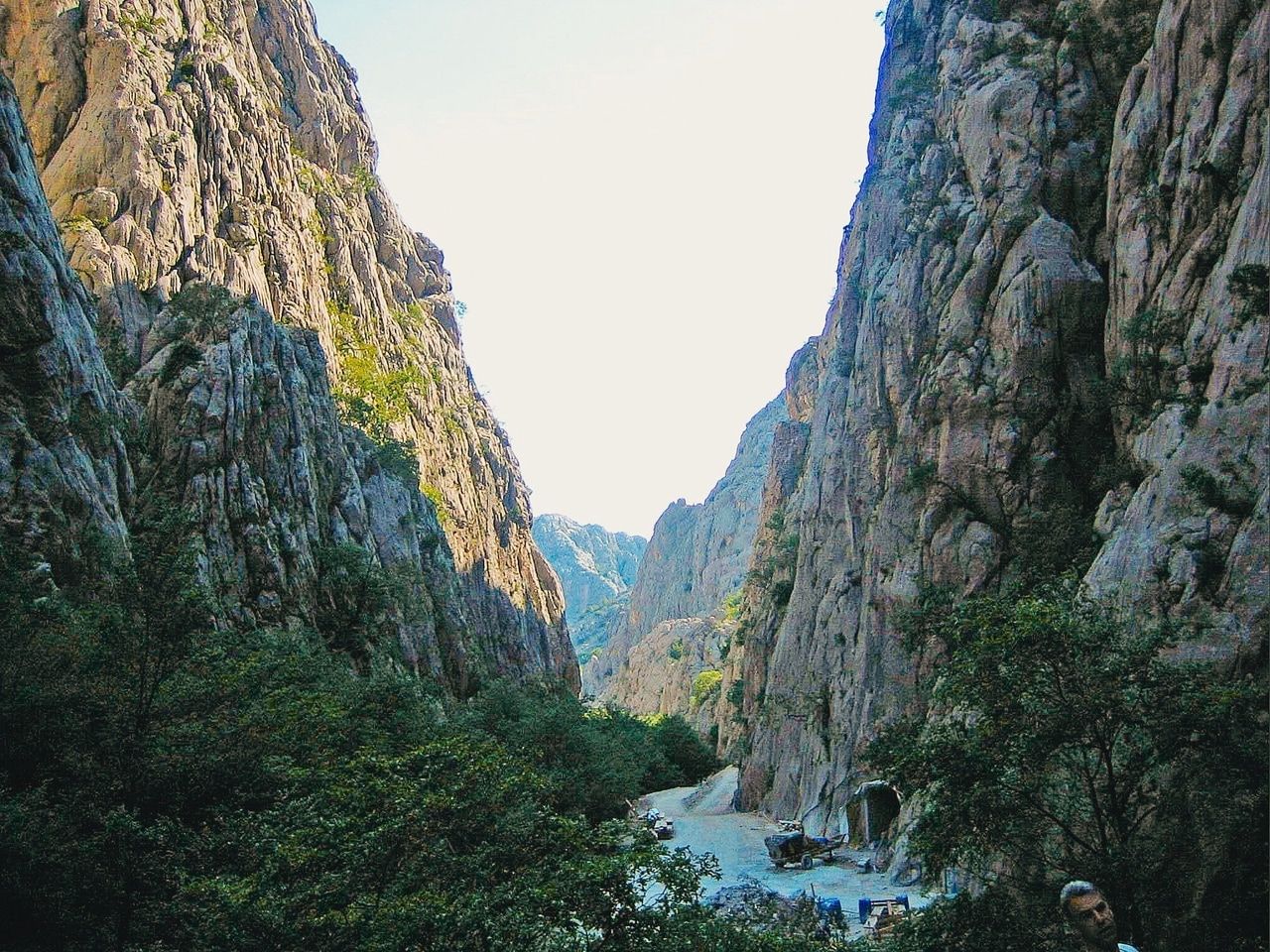 Gola rocciosa con sentiero stretto. Una gola impressionante con pareti rocciose ripide che si innalzano verso l'alto. Sul fondo della gola c'è un sentiero stretto, circondato da una vegetazione fitta. Sullo sfondo si vedono altre montagne che creano una formazione paesaggistica drammatica e naturale. Il cielo è sereno, il che mette in risalto le maestose formazioni rocciose e la profondità della gola.