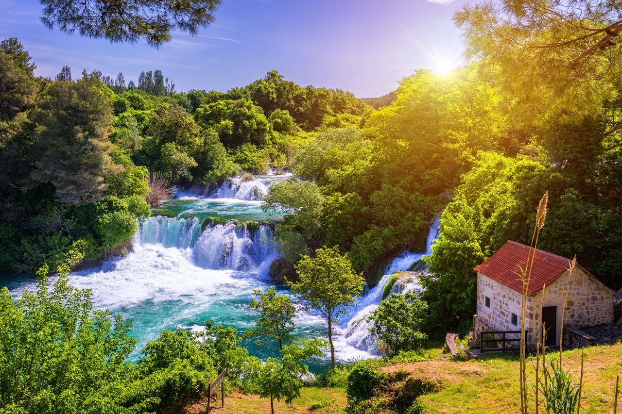 The waterfalls of Roški Slap in Krka National Park in Croatia. The water flows in several cascades through lush, green foliage and empties into a turquoise river. On the right side stands a small stone house with a red roof, surrounded by trees. The sun shines brightly, bathing the scene in a warm, golden light that highlights the natural beauty of the place.