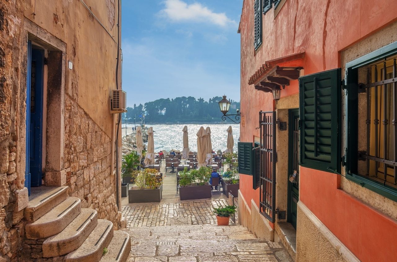 A narrow cobblestone path in Rovinj, Croatia, leads to a cozy outdoor area with tables and umbrellas right by the sea. On both sides of the path are historic buildings made of stone and colorful plaster with green shutters. In the background, the calm blue water stretches out, and trees and a small island can be seen on the horizon. The scene exudes a relaxed Mediterranean atmosphere.