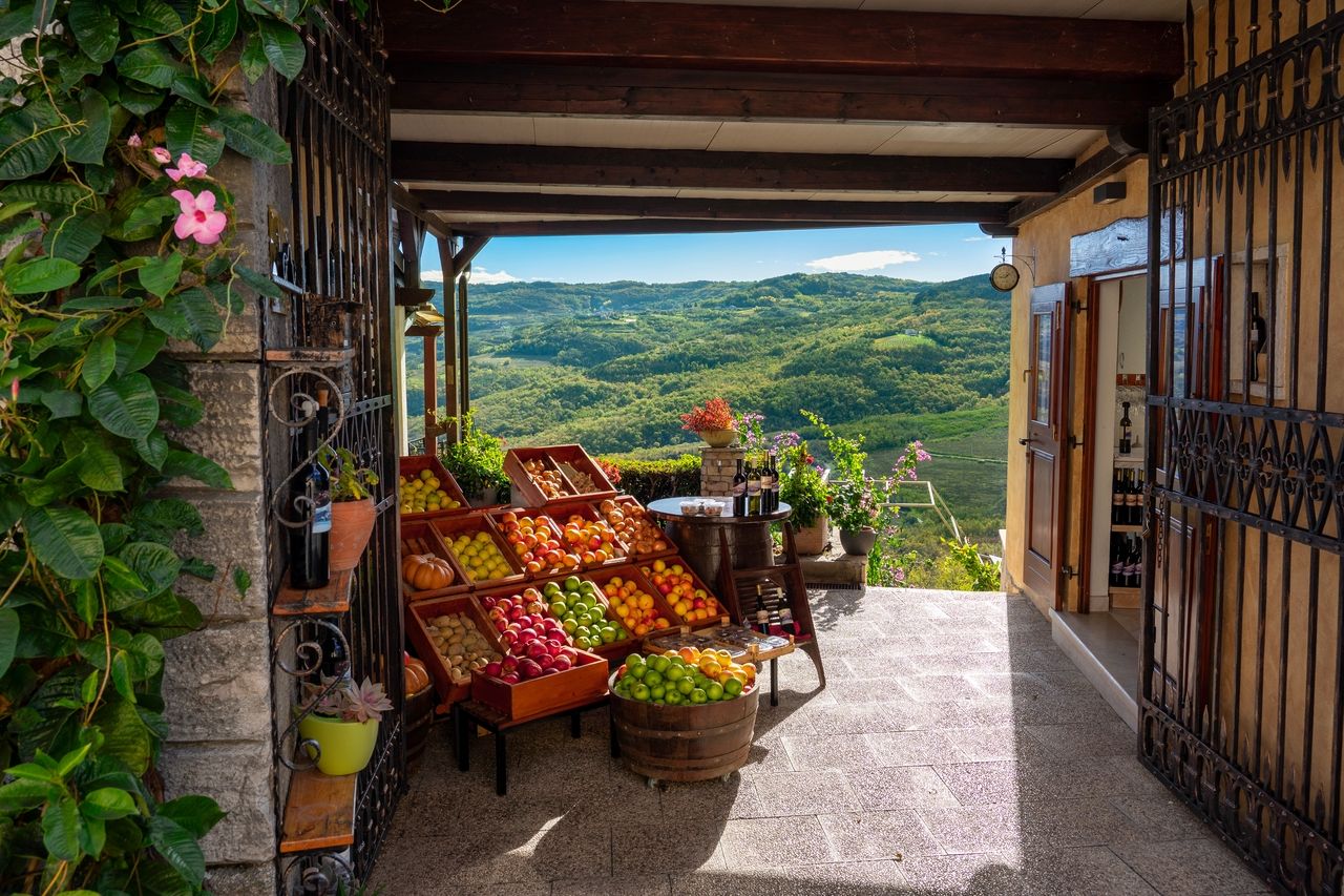 A rustic market stall in Motovun offering fresh fruits and vegetables with a picturesque view of the green hills and valleys in the background. The market is decorated with wooden crates, barrels, and wine bottles.