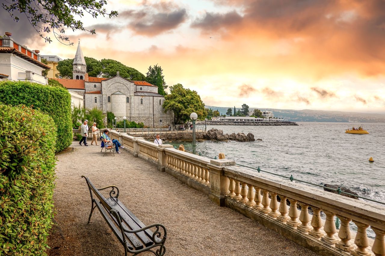 En malerisk spadseresti langs kysten i Opatija ved solnedgang. Folk nyder den afslappede atmosfære, sidder på bænke og går langs stien. En historisk kirke med markant arkitektur og røde tage ligger på den ene side, mens havet og kysten kan ses på den anden side.