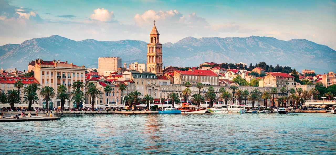 A photo of the coastline of Split with a view of Diocletian's Palace and the waterfront promenade. Palm trees line the promenade, behind which historic buildings with red-tiled roofs rise. In the background, the distinctive mountains provide an impressive backdrop for the city. Boats and yachts are anchored in the clear blue water of the harbor.