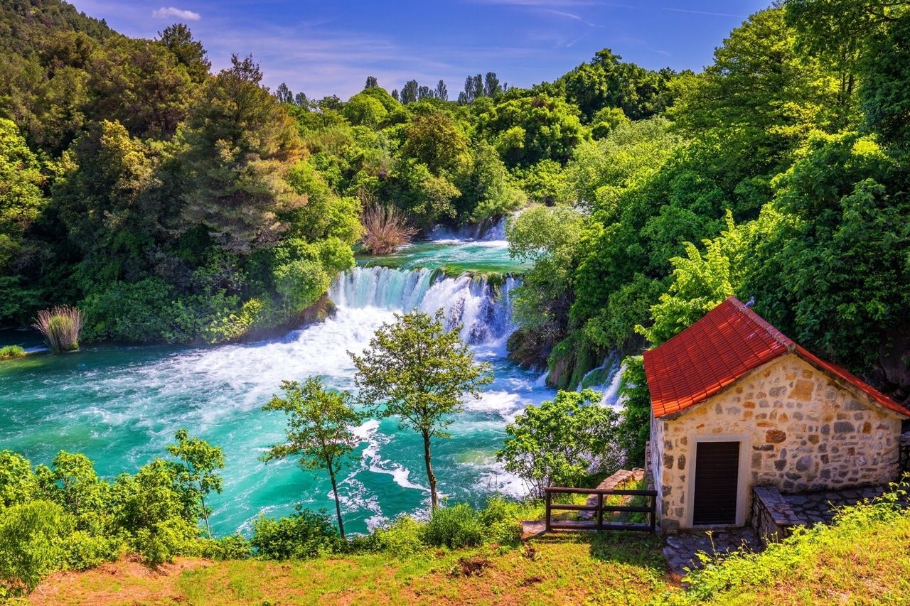 Şelale ve küçük taş ev ile pastoral bahar manzarası. Yemyeşil yapraklarla çevrili güzel bir şelale. Ön planda, kırmızı kiremit çatılı küçük bir taş ev, yeşil doğanın içine yerleştirilmiş. Açık mavi gökyüzü huzur ve doğa ile bağlantı hissi vererek sakin sahneyi tamamlıyor.