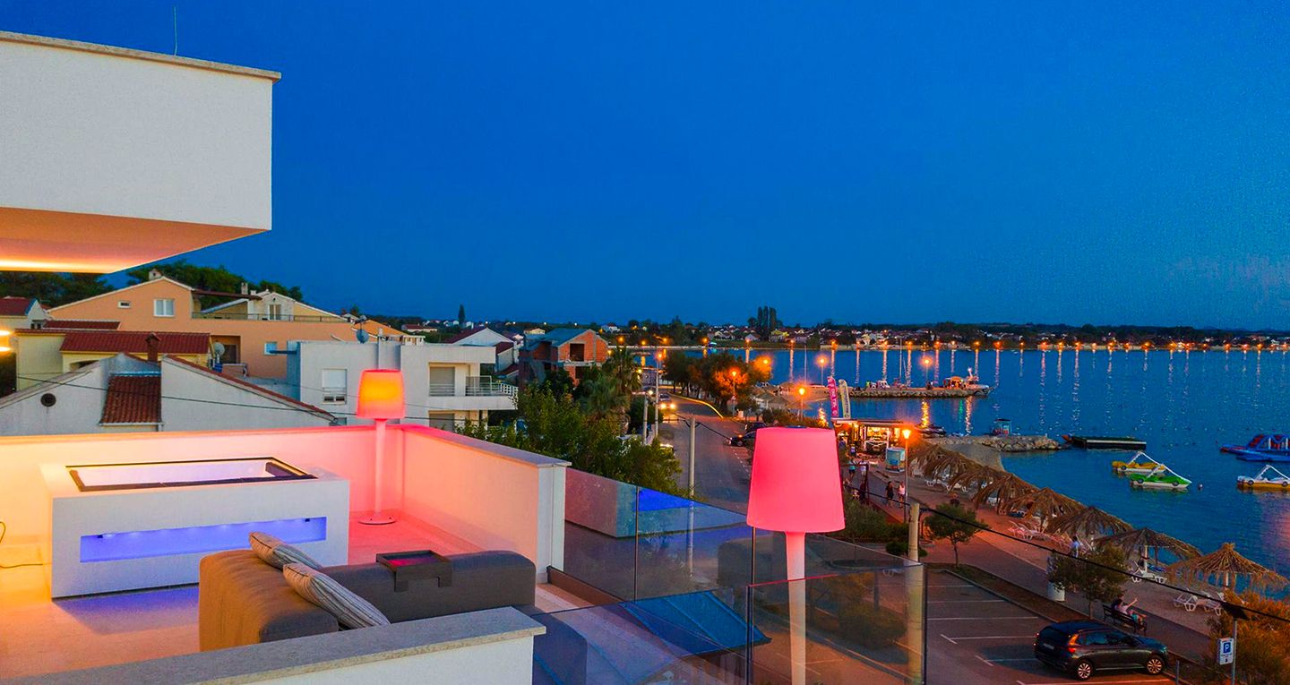 Feriebolig med havutsikt i Kroatia om natten. Moderne terrasse med stilfull belysning, utsikt over byen og havet med opplyste båter og strender. Ideell for en avslappende sommerferie.