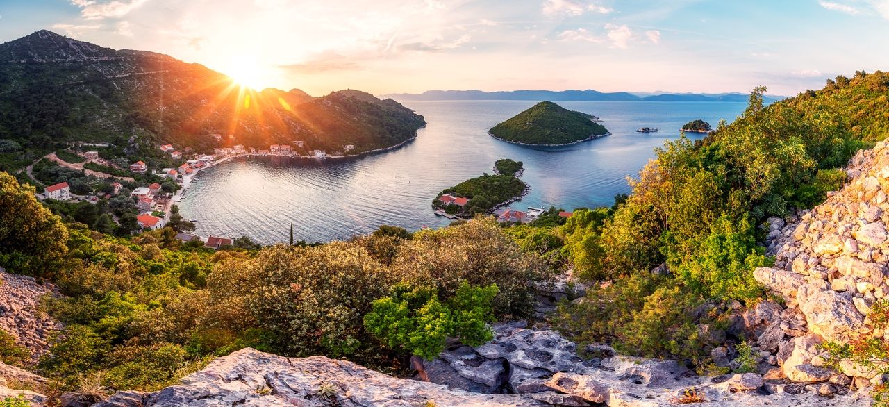 A breathtaking view of the island of Mljet at sunset. The sun sets behind the wooded hills, casting a golden light over the bay and the village. The calm water of the bay reflects the sky, and small islands can be seen in the background, surrounded by lush greenery and rocky slopes.