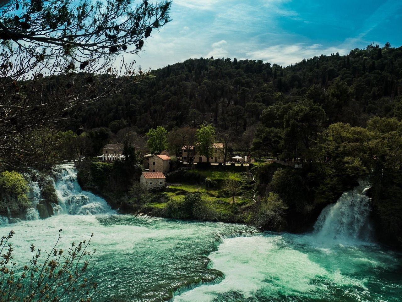 Pueblo con cascada en medio de un bosque. Un idílico pueblo con casas de piedra tradicionales se encuentra enclavado en un paisaje forestal verde. Dos impresionantes cascadas fluyen a través del pueblo, rodeadas de exuberante vegetación y altos árboles. El cielo está parcialmente nublado, lo que crea una atmósfera tranquila y natural.