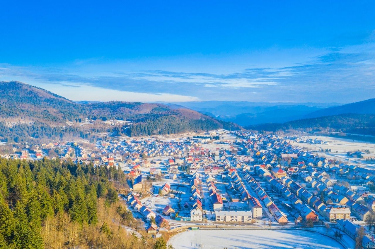 Vintervy av en liten stad i Kroatien, inbäddad i ett kuperat landskap och omgiven av snötäckta skogar. Den klara, kalla luften och den blå himlen ger scenen en lugn och idyllisk atmosfär.