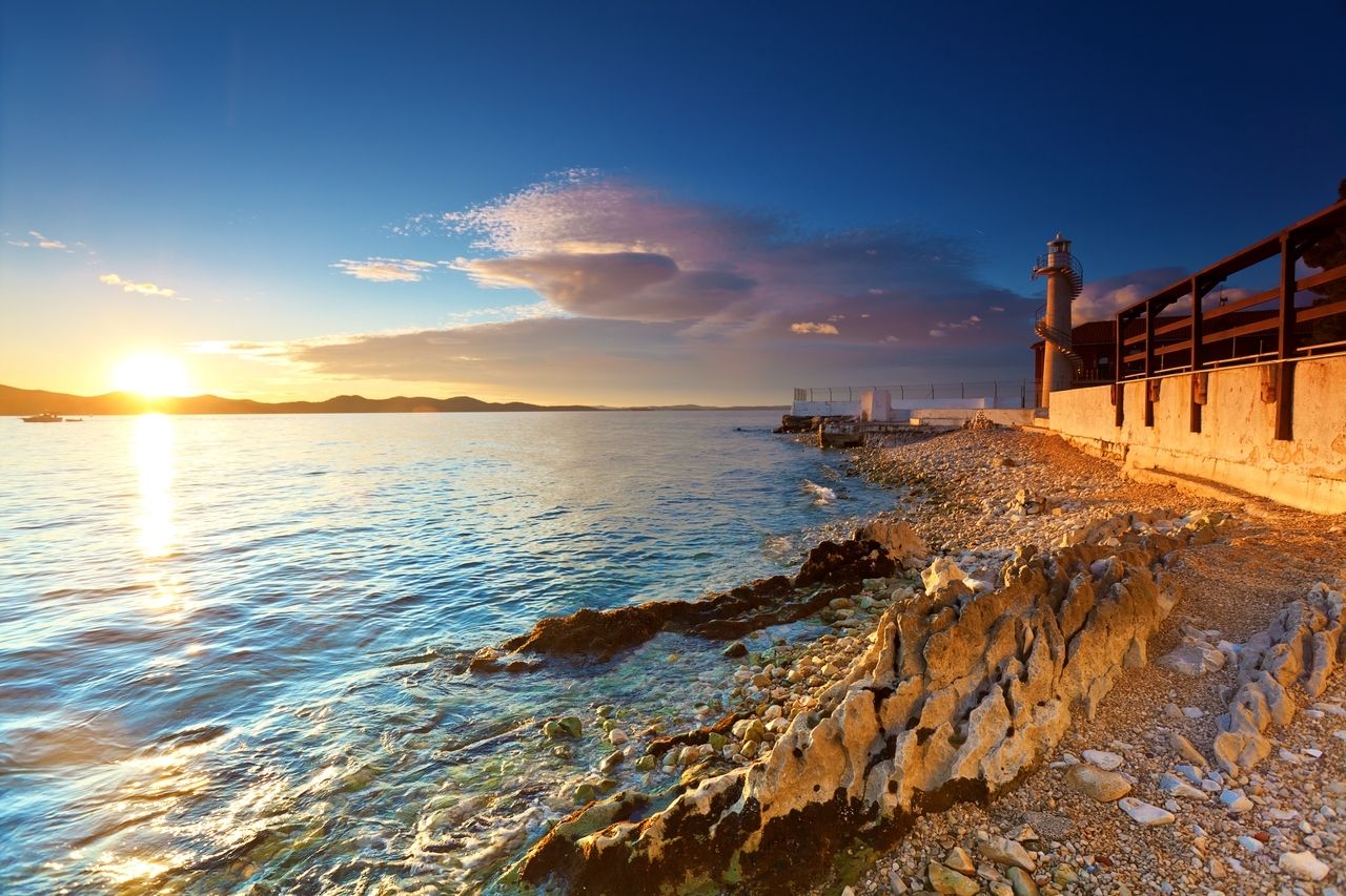 Sunset on the coast of Zadar, Croatia. The sun sets behind the mountains, bathing the sea in golden light. On the right edge of the picture is a lighthouse with a spiral staircase, standing on a rocky shore. The clear water shimmers in various shades of blue, and the scene exudes a calm and peaceful atmosphere.