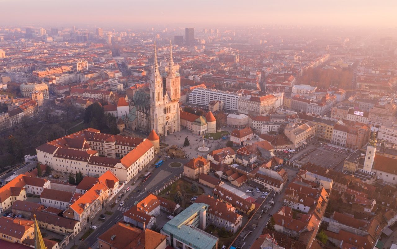 Flygfoto av Zagreb vid solnedgången med utsikt över Zagrebs katedral. Katedralen står i mitten av bilden och är omgiven av historiska byggnader med röda tak. Staden sträcker sig i fjärran och är badad i ett varmt, gyllene ljus som mjukt belyser byggnaderna och gatorna. Himlen är klar och staden verkar lugn och fridfull.