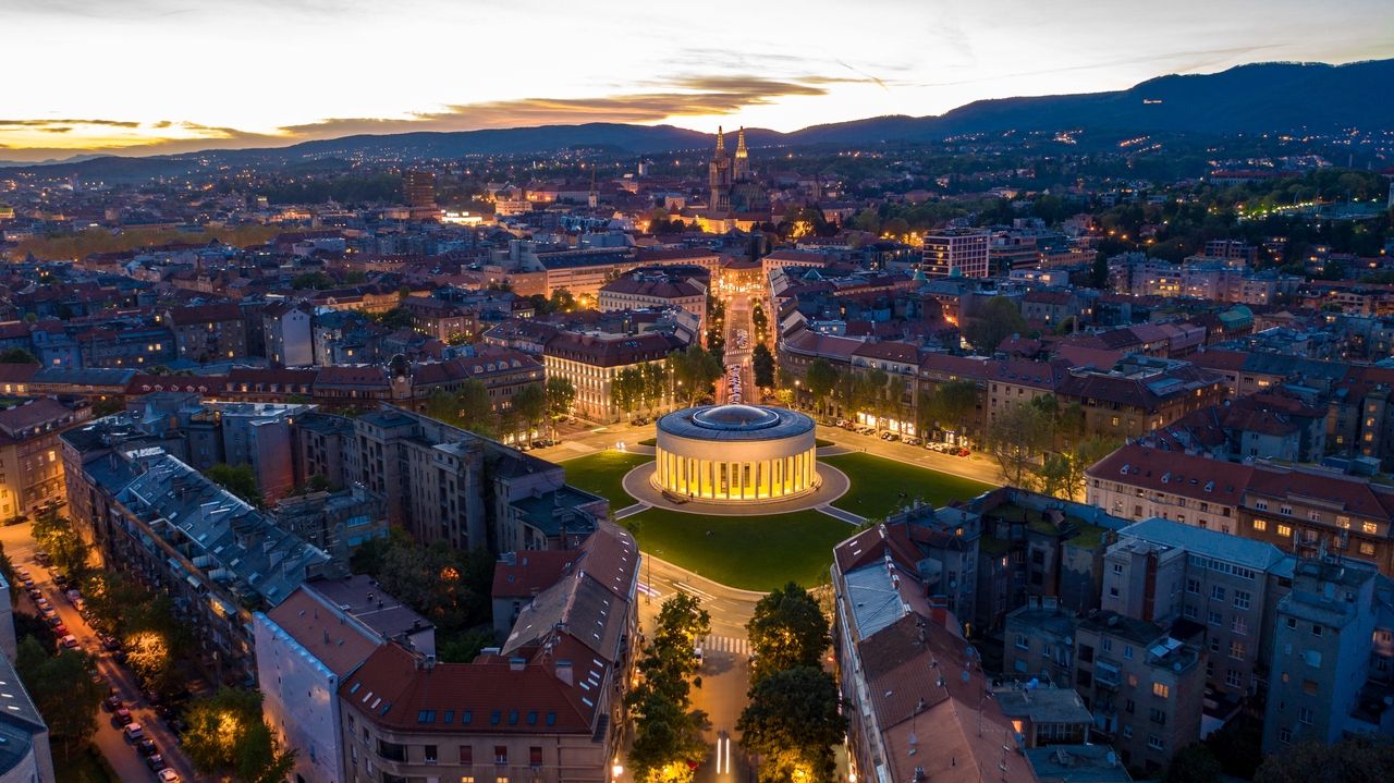 Aerial view of Zagreb at sunset. In the center of the image is the round illuminated building of the Croatian National Theater on a large square, surrounded by trees and illuminated streets. The city stretches into the background, with the distinctive silhouette of the Zagreb Cathedral in the distance. The mountains form a picturesque backdrop in the evening light.