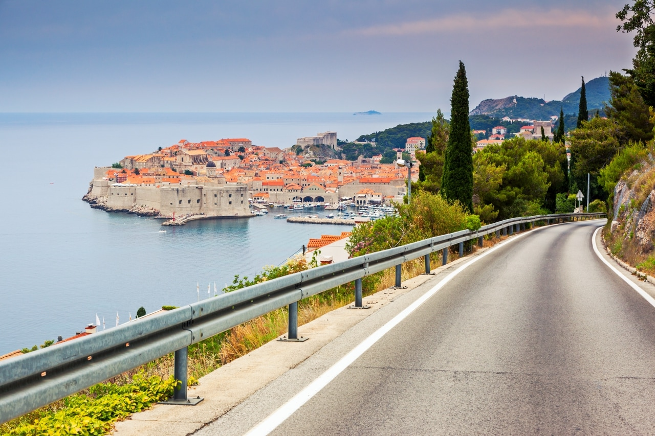 Panoramic view of the historic old town of Dubrovnik in Croatia, seen from a winding road leading down to the coast. The ancient city walls and red roofs contrast with the clear blue sea and the surrounding lush vegetation.