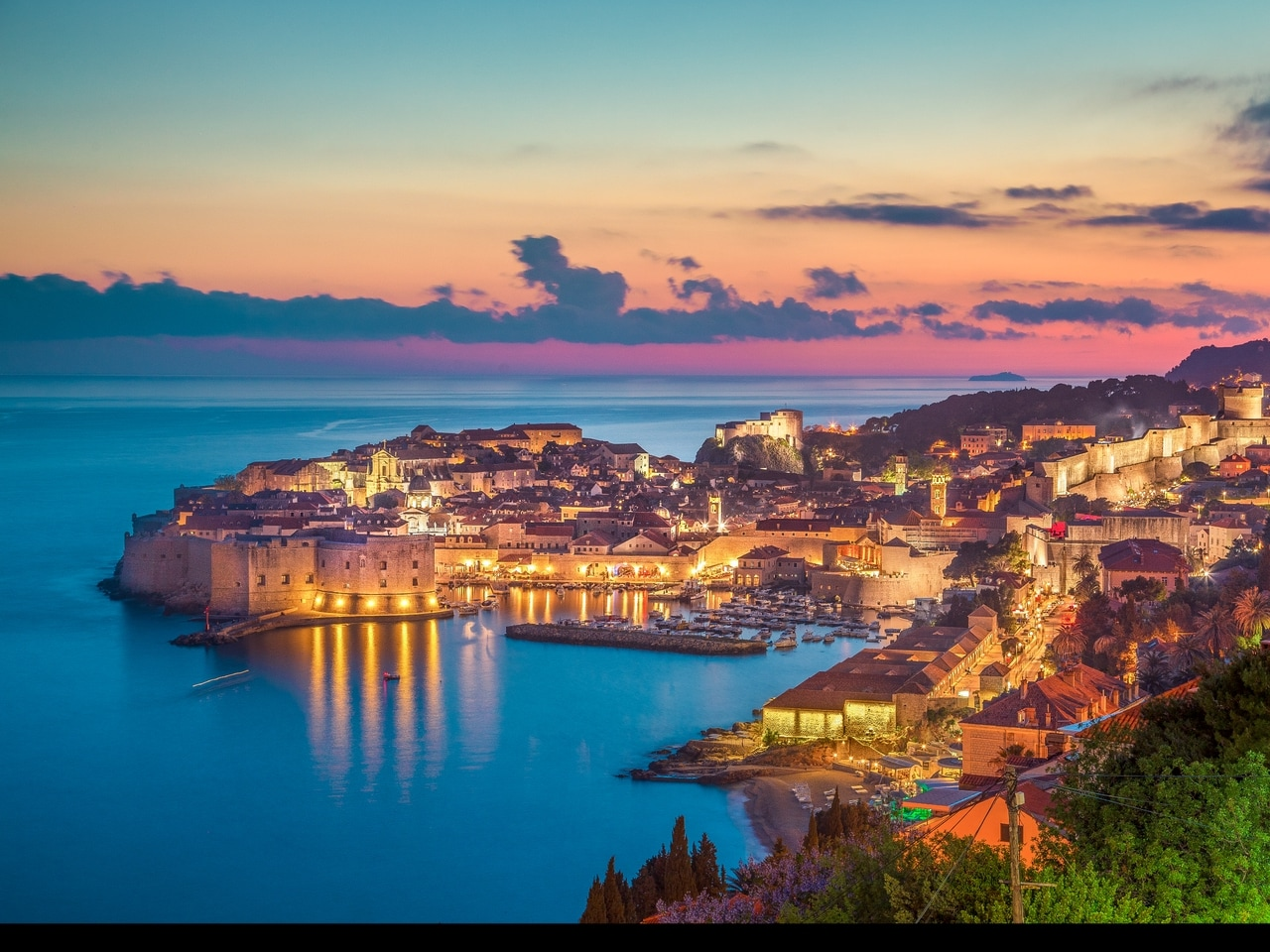 Oplyst gamle bydel i Dubrovnik i Kroatien ved solnedgang. De historiske bymure og bygninger spejler sig i det rolige Adriaterhav og skaber en romantisk og malerisk atmosfære.