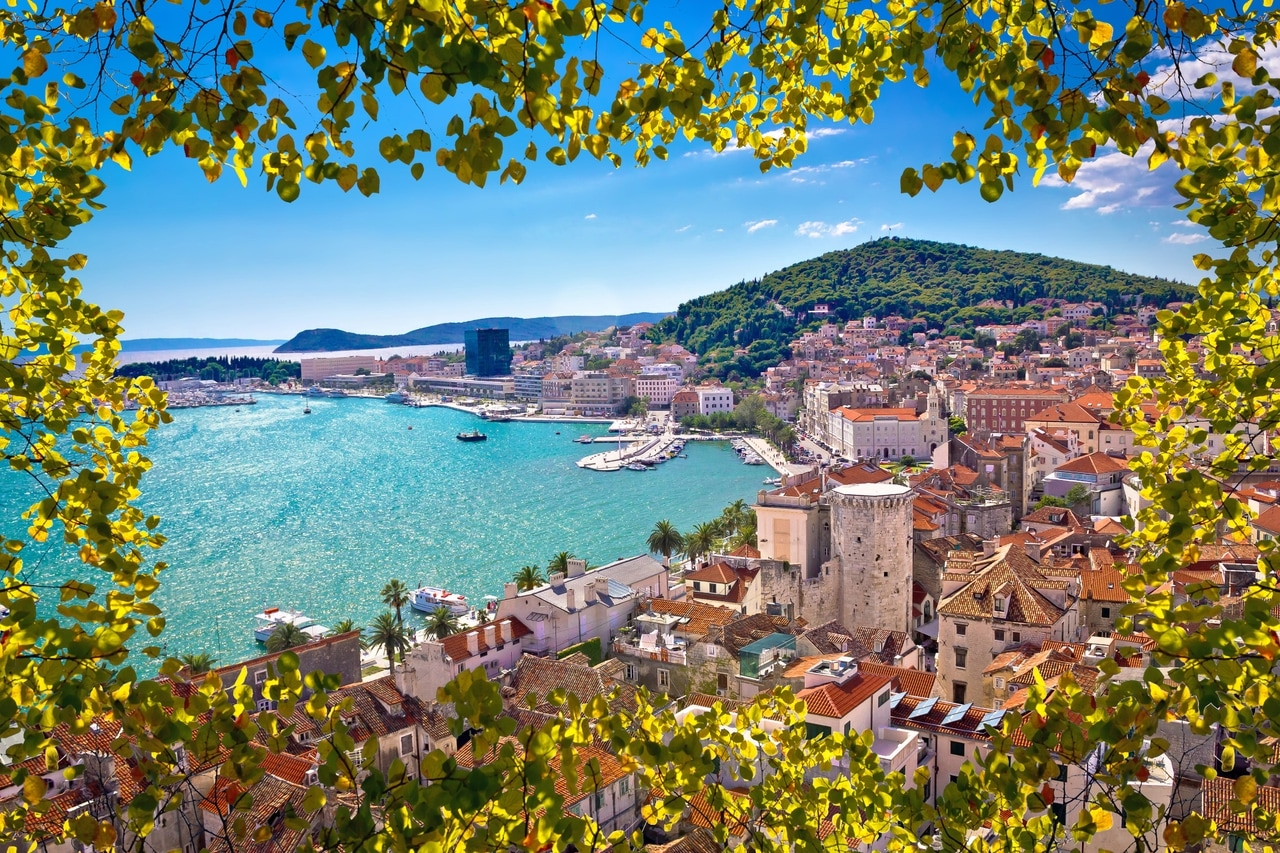 Panoramablik over kystbyen Split i Kroatien, indrammet af grønne blade. De historiske bygninger, den maleriske havn og de omkringliggende bakker tilbyder et betagende landskabsbillede under en klar, blå himmel.