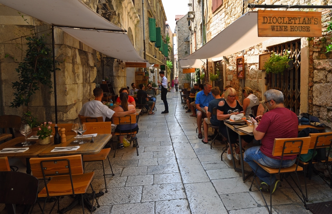 Livlig gatubild i gamla stan i Split, Kroatien, med människor som sitter på en mysig utomhusrestaurang. De smala, stenlagda gränderna och de historiska byggnaderna skapar en charmig och inbjudande atmosfär.